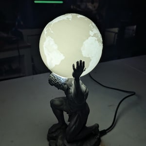 Könnte beinhalten: Eine dunkelgraue, kniende Figurenskulptur, die eine leuchtende, kugelförmige Lampe in Form der Erde trägt. Der Globus zeigt beleuchtete weiße Kontinente auf einer sanft leuchtenden Oberfläche, während die Figur in einer muskulösen, kauernden Haltung auf einem felsigen Sockel dargestellt ist und mit einem schwarzen Stromkabel verbunden ist.