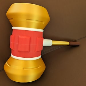 May include: A gold and red toy hammer with a white and gold handle. The hammer has a rounded head with a red, textured center.