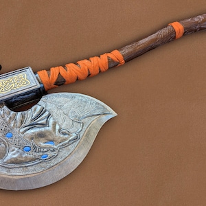 Puede incluir: Un hacha de madera con un mango tallado y una cabeza de metal. La cabeza está decorada con un diseño de dragón y tiene un borde dorado. El mango está envuelto en un cordón naranja.