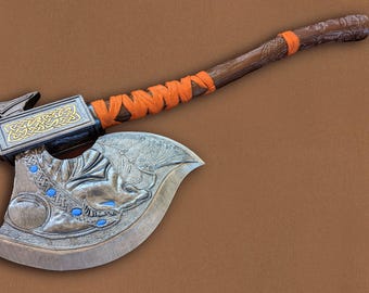 May include: A wooden axe with a carved handle and a metal head. The head is decorated with a dragon design and has a gold trim. The handle is wrapped in orange cord.
