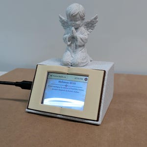 May include: A stone-textured angel figurine kneeling in prayer atop a rectangular base shaped like an open book. The book features a digital screen displaying the text "Hebreos 10:15" and "Y nos atestigua lo mismo el Espíritu Santo; porque después de haber dicho".