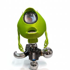 Puede incluir: Una figura verde, similar a un dibujo animado, con un ojo grande, brazos y boca, montada sobre un enganche de remolque negro y plateado. El enganche tiene el texto "REESE TOWPOWER". La figura es un objeto decorativo y caprichoso.