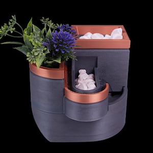 Op de afbeelding: Een drielaagse plantenbak in grijs en koper met een kleine plant en witte stenen. De plantenbak is gemaakt van een getextureerd materiaal.