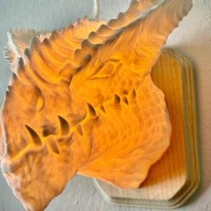 May include: A detailed, illuminated dragon head sculpture mounted on a wooden plaque. The dragon head is a light beige color with intricate textures and sharp teeth. The light source creates a warm glow, highlighting the details of the sculpture.