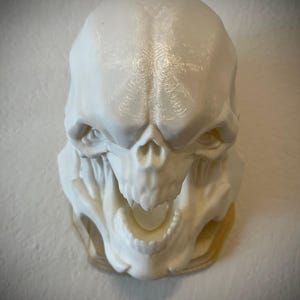 Puede incluir: Un cráneo blanco impreso en 3D con una gran boca abierta y dientes afilados. El cráneo está detallado con una superficie texturizada y parece ser un objeto decorativo.