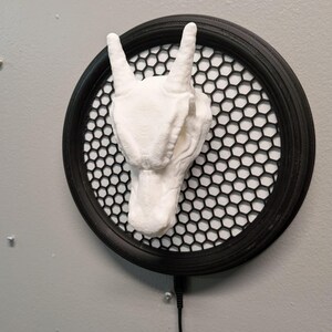Puede incluir: Una escultura de mano blanca impresa en 3D en el gesto de 'rock on', montada sobre un marco negro redondo con una malla de panal. La escultura está fijada a una pared gris con un cable negro.