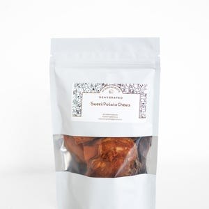 May include: A white pouch with a clear window containing dehydrated sweet potato chews. The pouch has a label with the text "Sweet Potato Chews" and a border of dog illustrations. The product is for pets.