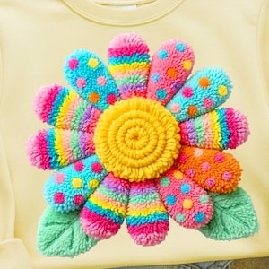 May include: A light yellow sweatshirt featuring a large, colorful flower design. The flower has a yellow center and vibrant petals in shades of pink, blue, orange, and green. The petals are decorated with polka dots and rainbow stripes.