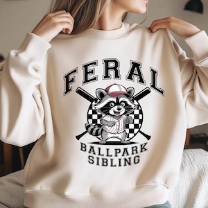 以下が含まれることがあります： クリーム色のスウェットシャツに、野球帽をかぶり、バットを持ったアライグマのグラフィックがプリントされています。アライグマの下には「FERAL BALLPARK SIBLING」というテキストがプリントされています。