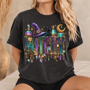 May include: A black t-shirt featuring a graphic print with the word WITCHY in large, ornate, metallic-toned letters. The design includes a purple witch hat, a black cat, glowing potion bottles, crystals, and a golden crescent moon.