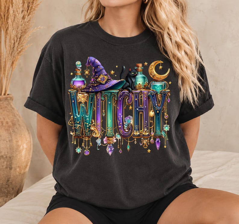 May include: A black t-shirt featuring a graphic print with the word WITCHY in large, ornate, metallic-toned letters. The design includes a purple witch hat, a black cat, glowing potion bottles, crystals, and a golden crescent moon.