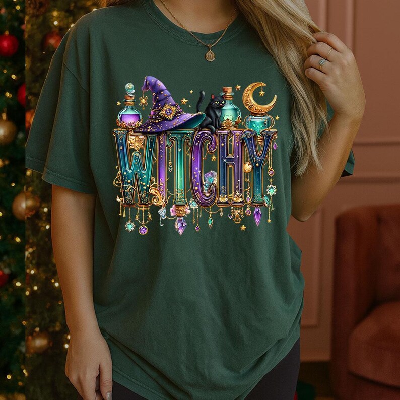 May include: A dark green t-shirt featuring a graphic print of the word WITCHY in large, colourful, ornate letters. The design includes a purple witch hat, a black cat, glowing potion bottles, a crescent moon, and various hanging crystal charms.