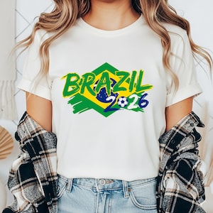 以下が含まれることがあります： 白いTシャツ。緑色の筆記体で「BRAZIL」の文字、緑と黄色のひし形模様、サッカーボールのグラフィック。数字の「2026」は青色。Tシャツは人が着ています。