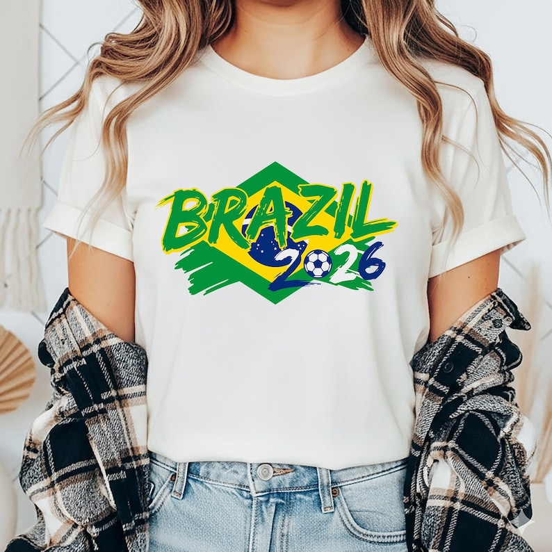 以下が含まれることがあります： 白いTシャツ。緑色の筆記体で「BRAZIL」の文字、緑と黄色のひし形模様、サッカーボールのグラフィック。数字の「2026」は青色。Tシャツは人が着ています。