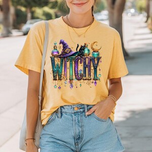 May include: A mustard yellow t-shirt featuring a graphic print with the word WITCHY in large, ornate, multi-coloured letters. The design includes a purple witch hat, a black cat, glass potion bottles, and a golden crescent moon with hanging crystals.