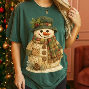 May include: A dark green t-shirt featuring a whimsical snowman design. The snowman wears a green hat, a plaid scarf, and has button eyes and a carrot nose. The snowman is made of patchwork fabric and has a pinecone at its base.