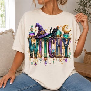 May include: A cream-coloured t-shirt featuring a graphic print with the word WITCHY in large, ornate, gradient-coloured letters. The design includes a purple witch hat, a black cat, glass potion bottles, a golden crescent moon, and hanging crystals.