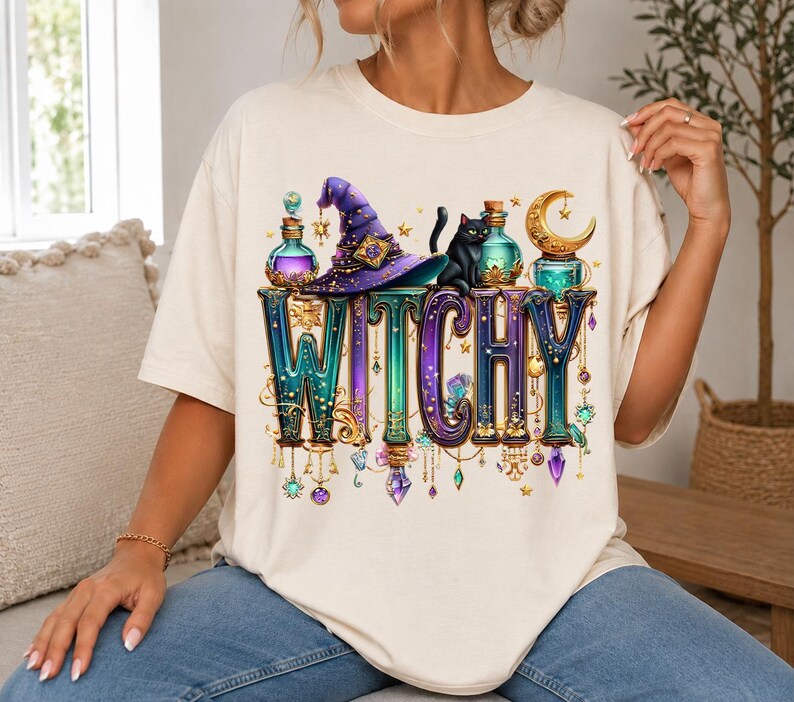 May include: A cream-coloured t-shirt featuring a graphic print with the word WITCHY in large, ornate, gradient-coloured letters. The design includes a purple witch hat, a black cat, glass potion bottles, a golden crescent moon, and hanging crystals.