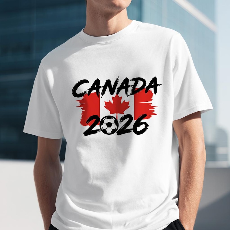 以下が含まれることがあります： 白いTシャツ。黒い筆記体で「CANADA」の文字が書かれ、その上に赤と白のカナダ国旗のデザイン。旗の下にはサッカーボールのグラフィックと数字の「2026」が描かれています。