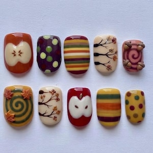 May include: A set of ten press-on nails featuring autumn-themed designs including apple slices, colorful horizontal stripes, polka dots, bare tree branches with small leaves, swirling patterns with orange maple leaves, and three-dimensional acorns.