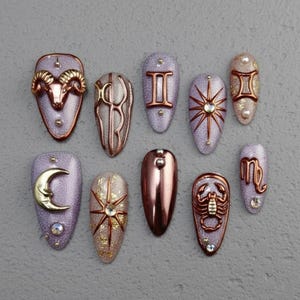 May include: A collection of ten artificial nails with various zodiac-themed designs. The nails feature metallic accents in copper and gold, with symbols like a ram, sun, moon, and scorpion. The base colors include glittery lavender, gold, and copper.