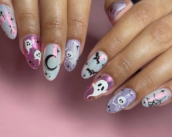 Może przedstawiać: Owalny manicure z motywami Halloween. Paznokcie zdobią duchy, nietoperze, pająki, pajęczyny, księżyce i czaszki w kolorach białym, fioletowym i różowym, na różowym tle.