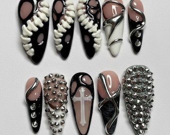 May include: A set of ten long, coffin-shaped artificial nails. Designs include black and white skeleton-like patterns, silver accents, and studded details. Some nails feature a nude base with silver or black embellishments.