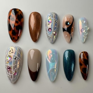 May include: An assortment of ten almond-shaped artificial nails. The nails feature various designs, including tortoiseshell, glitter, iridescent, and geometric patterns. Some nails are adorned with rhinestones and other embellishments. The color palette includes brown, beige, and teal.