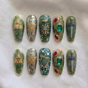 May include: A collection of ten artificial nails with detailed designs. The nails showcase gold and silver accents, such as a Triforce symbol, a Hylian Crest, and a sword. Some nails have green and pink backgrounds with gemstones.