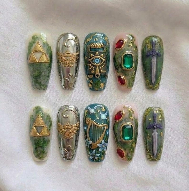 May include: A collection of ten artificial nails with detailed designs. The nails showcase gold and silver accents, such as a Triforce symbol, a Hylian Crest, and a sword. Some nails have green and pink backgrounds with gemstones.