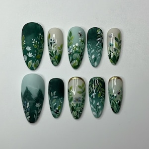 Op de afbeelding: Een set van tien kunstnagels met een op de natuur geïnspireerd ontwerp. Elke nagel heeft een ander ontwerp, waaronder groene bladeren, witte bloemen en kleine insecten, allemaal geschilderd op een lichtgroene of witte basis. Sommige nagels hebben een gouden rand.