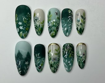 May include: A set of ten artificial nails with a nature-inspired design. Each nail features a different design, including green leaves, white flowers, and small insects, all painted on a light green or white base. Some nails have a gold trim.