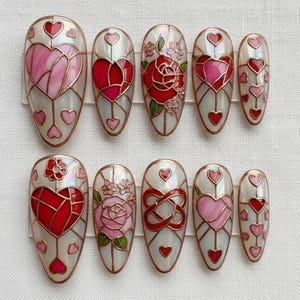 Puede incluir: Un conjunto de diez u&ntilde;as artificiales con un dise&ntilde;o de vidriera. Las u&ntilde;as presentan patrones de corazones y flores en rojo, rosa y blanco, delineados con un borde color cobre. Algunas u&ntilde;as tienen peque&ntilde;os acentos de coraz&oacute;n.