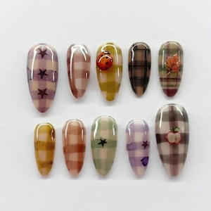 Può includere: Un set di dieci unghie finte a mandorla con vari motivi a quadri in tonalità viola, marrone, giallo, verde e beige. Diverse unghie sono decorate con piccoli ornamenti 3D tra cui una coccinella, una foglia autunnale e una mela.
