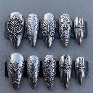 May include: A collection of ten silver-toned nail covers. Designs include a rose, swirling patterns, a butterfly, a star, and daggers. The nail covers are arranged on a dark gray surface.