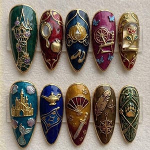 May include: A set of ten almond-shaped press-on nails featuring intricate gold-toned metal relief designs on colorful enamel backgrounds. Each nail displays unique motifs including a tower, mirror, apple, carriage, spinning wheel, rose, castle, lamp, fan, sword, compass, and bear paw.