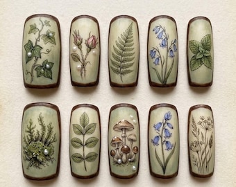 May include: A collection of ten rectangular, hand-painted wooden pieces with botanical designs. Each piece features a different plant, including ivy, roses, ferns, bluebells, and mushrooms, with a pearl accent. The pieces have a light green background and a dark brown border.