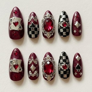 May include: A set of ten artificial nails in various designs. The nails feature a deep red glitter base, with accents including playing card motifs, checkerboard patterns, and ornate silver embellishments with red gems. The nails are oval-shaped.