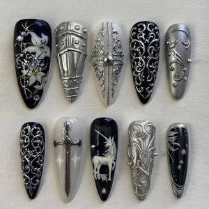 May include: A set of ten almond-shaped press-on nails featuring intricate designs in black, white, and metallic silver. The collection includes motifs such as white lilies, silver armor plating, ornate filigree, a silver sword, and a white unicorn.