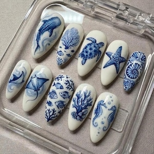 Peut inclure: Dix faux ongles ovales blancs, chacun peint à la main avec un motif marin bleu différent. Les motifs comprennent une baleine, du corail, une tortue, une étoile de mer et un hippocampe. Les ongles sont présentés dans un récipient en plastique transparent.