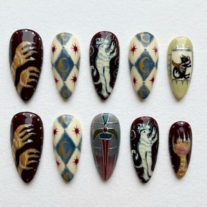 Peut inclure: Dix faux ongles en forme d'amande avec des motifs complexes. Les ongles présentent divers motifs, dont un chat, une main griffue et des formes géométriques dans des tons bordeaux, crème, bleu et or.