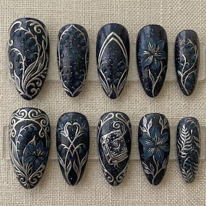 May include: A set of ten dark blue press-on nails with silver floral and geometric designs. Some nails feature a 3D flower, while others have a book design. The nails are oval-shaped and have a glittery finish.
