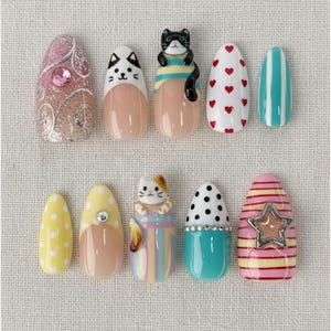 May include: A collection of ten artificial nails with various designs. The nails feature patterns like polka dots, stripes, hearts, and cat-themed embellishments. Colors include pink, yellow, white, and blue, with some nails adorned with rhinestones.