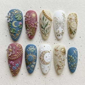 May include: A collection of ten almond-shaped, painted artificial nails. Each nail features intricate designs, including flowers, moons, suns, and butterflies, with gold accents. Colors include blue, pink, white, and beige, creating a whimsical, artistic aesthetic.