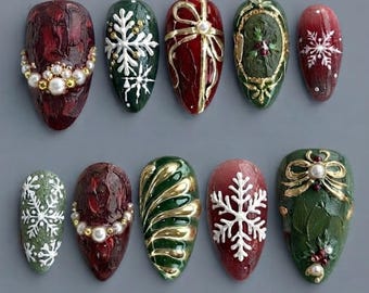 May include: A set of ten almond-shaped press-on nails featuring festive holiday designs. The collection includes deep red and forest green bases decorated with white 3D snowflakes, gold ribbon accents, pearl embellishments, and ornate frames.