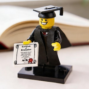 May include: A Lego figurine of a graduate wearing a black cap and gown, holding a certificate that reads "Certificate of Graduation." The figurine has a yellow head and is standing on a black base. An open book is in the background.