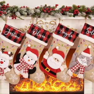 May include: Four Christmas stockings hang above a fireplace, each with a name: Justine, Olivia, Angela, and Selina. The stockings have a red and black plaid cuff and are decorated with festive characters like a snowman, penguin, Santa, and a cat.
