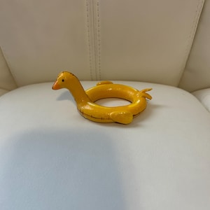 May include: A yellow, ring-shaped toy resembling the Loch Ness Monster. It features a long neck, small head with an orange beak, and a yellow ring body with small fins. The toy is placed on a white surface.