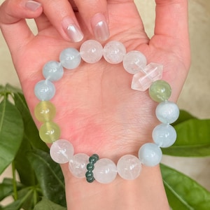 May include: A beaded bracelet featuring a mix of round and faceted gemstones in shades of white, light blue, green, and clear. The bracelet is held up against a light background, showcasing the various colors and textures of the stones.