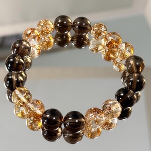 Könnte beinhalten: Ein Armband mit abwechselnd dunkelbraunen und goldbraunen Kristallperlen. Das Armband wird auf einer reflektierenden Oberfläche präsentiert, die die runden Perlen und ihre Farbvariationen zeigt. Ein Modeaccessoire.